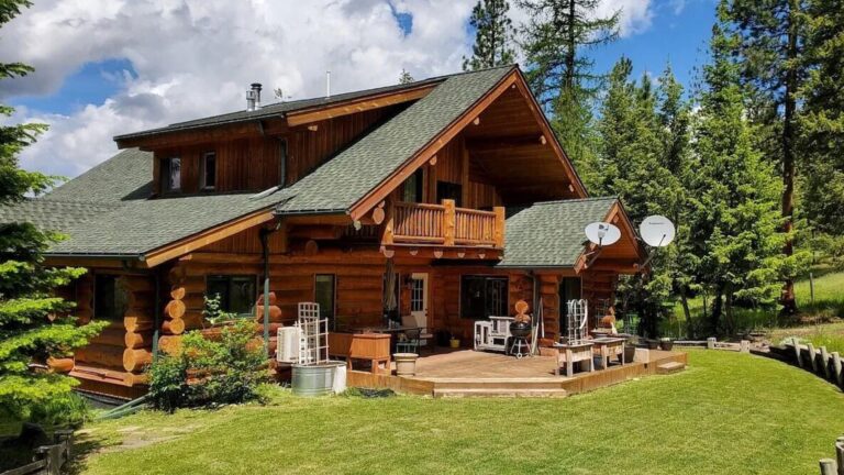 log cabin with green roof with a big deck around the house, surrounded by pine trees and greenery