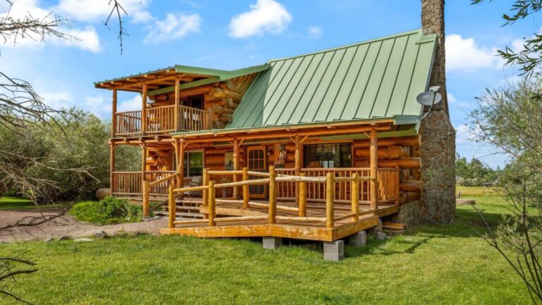 exterior of a log cabin with beautiful log work on the walls and the banisters around the poch, slanted metal green roof, a balcony, and greenery around the cabin