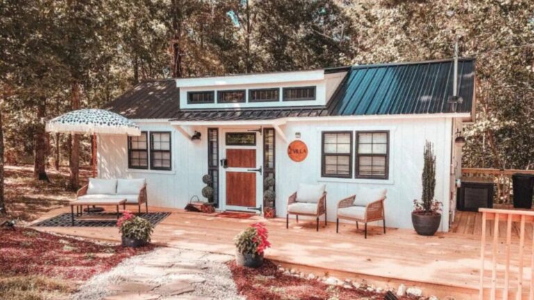 exterior of a tiny home with a porch with white cushioned chairs and a parasol set in the woods