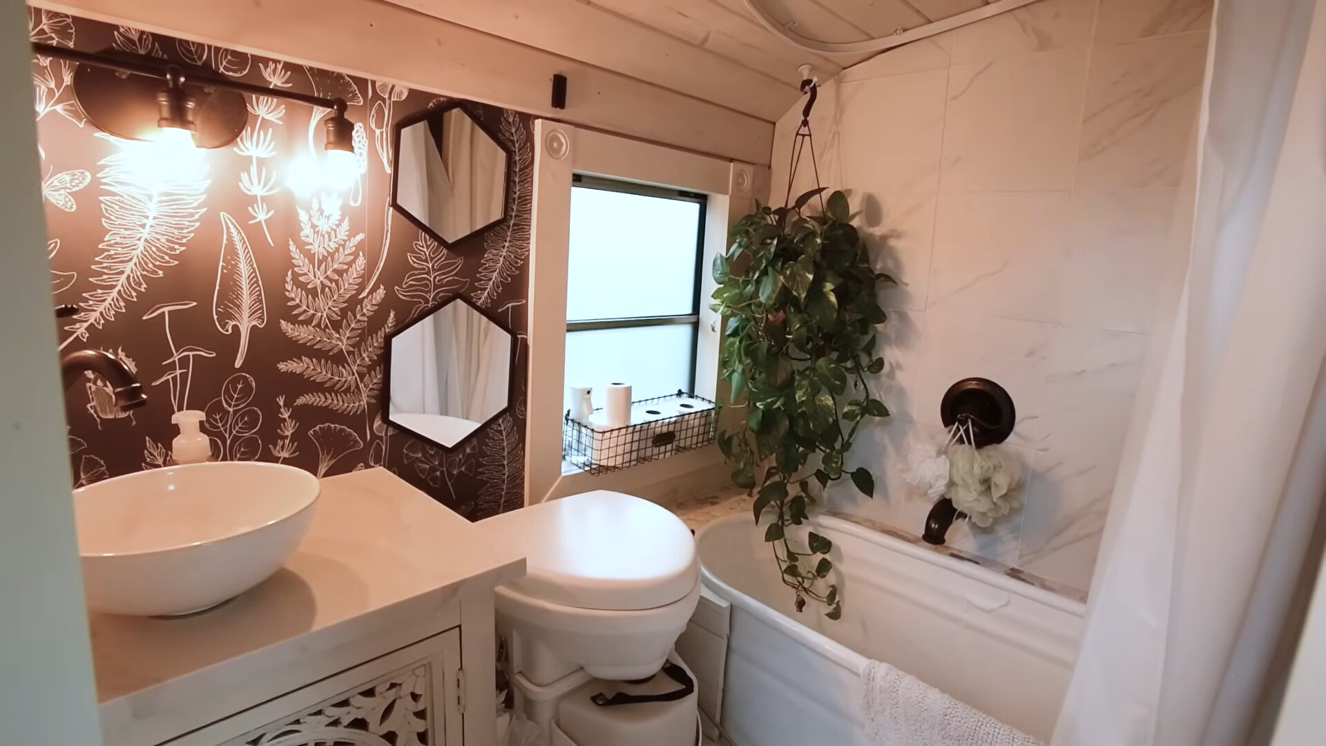 bathroom with a white bathtub, white sink on a white countertop, white toilet and white tiles, one brown wall with plant textures