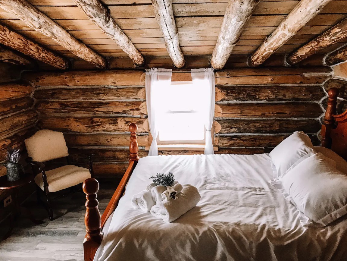 bedroom with a big bed with white bedsheets, chair in the corner with a tiny brown table, a window with white curtains and brown, wooden walls