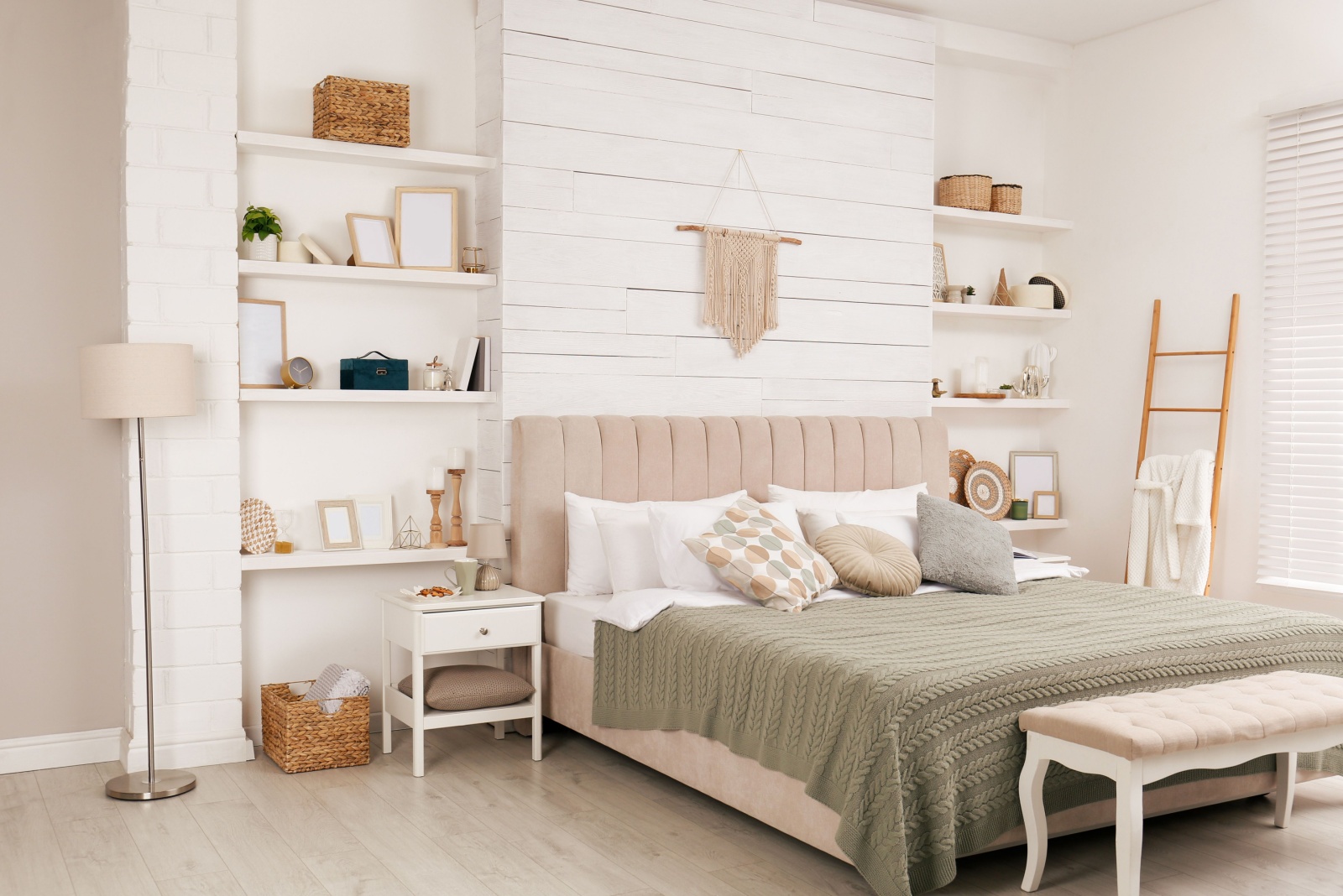 bedroom with a comfortable bed, a cushioned bench in front of the bed, built-in white shelves filled with photo frames and other accessories and a tiny white bedside table