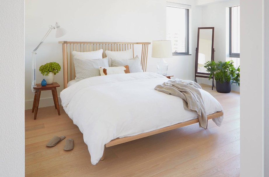 bedroom with a bed with white bed sheets, wooden bed frames, brown bedside table with a plant and a lamp, a full body mirror in the corner