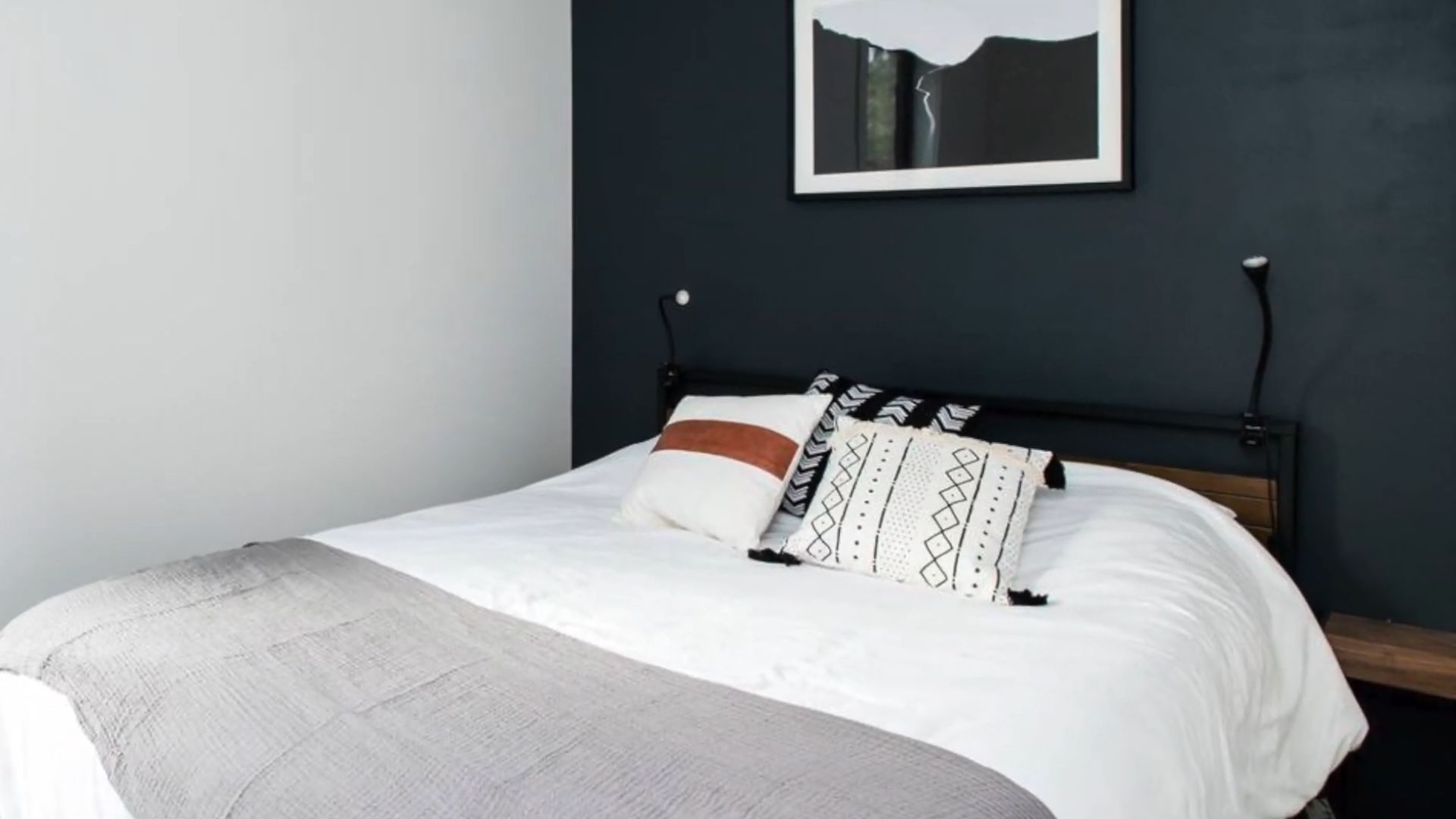 simple bedroom with a cozy bed and a grey blanket over it, leaning onto the black accent wall with a picture on it