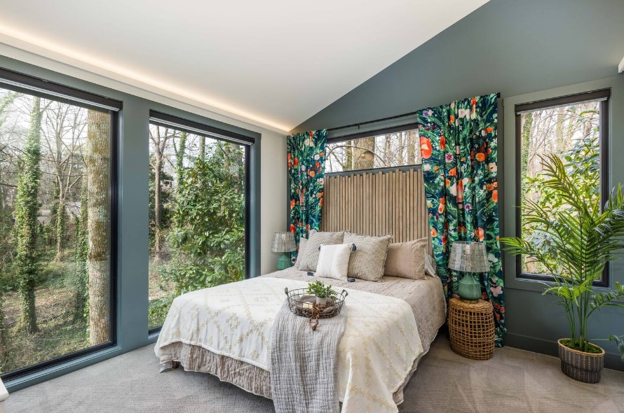 bedroom with green and white walls, floral patterned curtains, a bed, floor to ceiling windows with the view of nature and a big plant