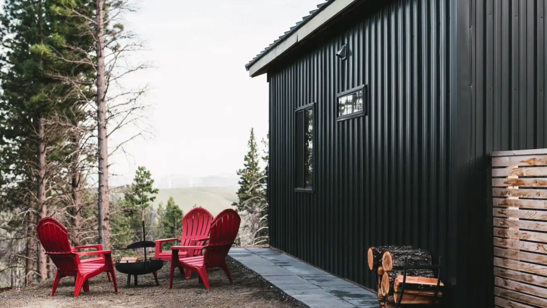 exterior of the black cabin with wood siding, fire pit area on the left with bright red chairs