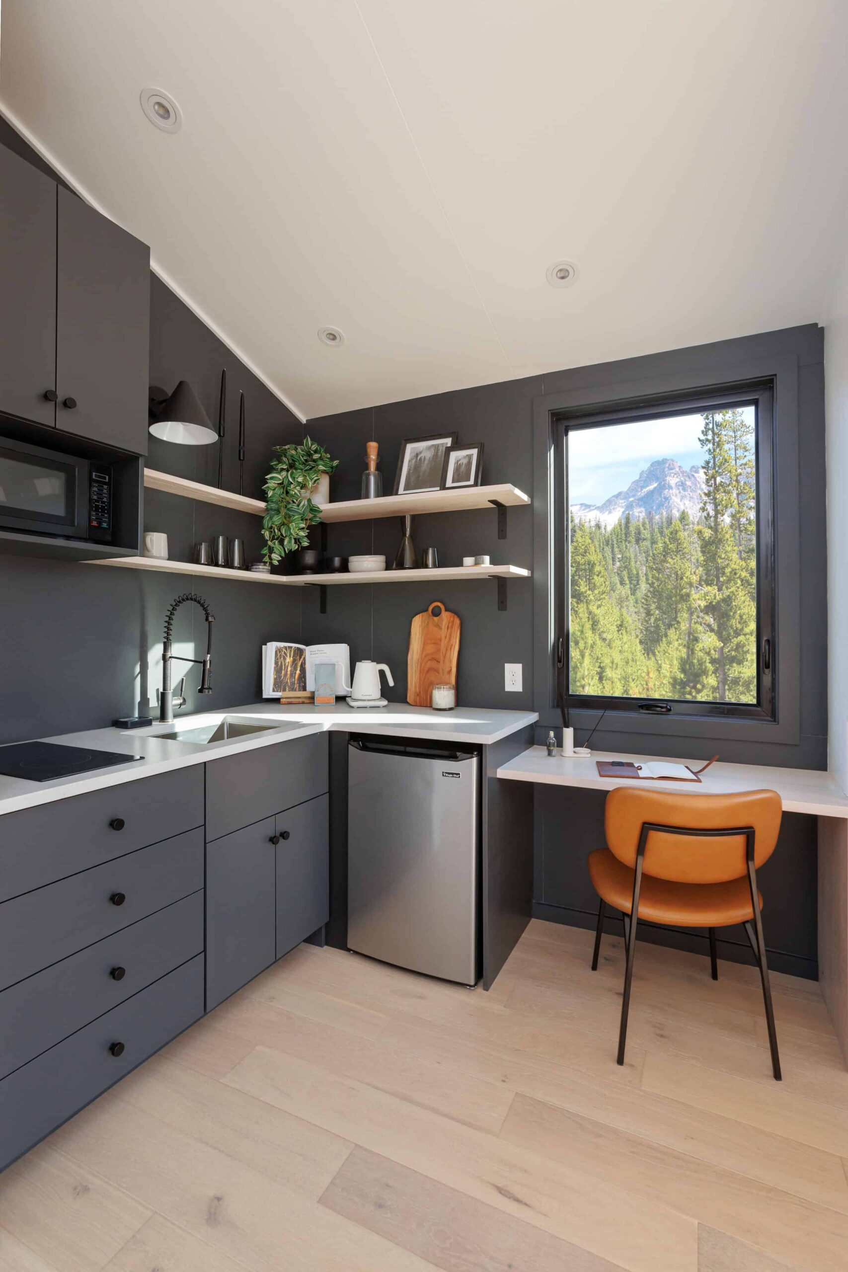 kitchen with dark gray walls, dark gray cabinets, white countertop, white dining table, orange chair, window above the able and shelves