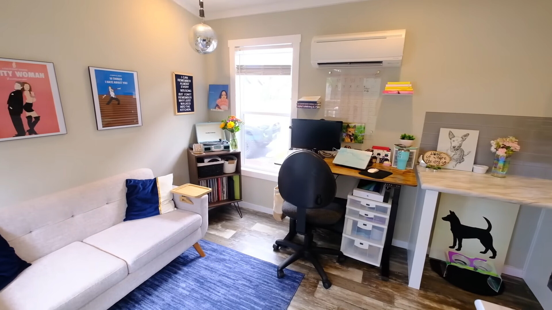 living room with a white couch, movie posters framed above the couch, brown cabinet in the corner with open shelves filled with vinyls and a vinyl record on top, office area with a brown desk and a black chair, and a floating shelf with books