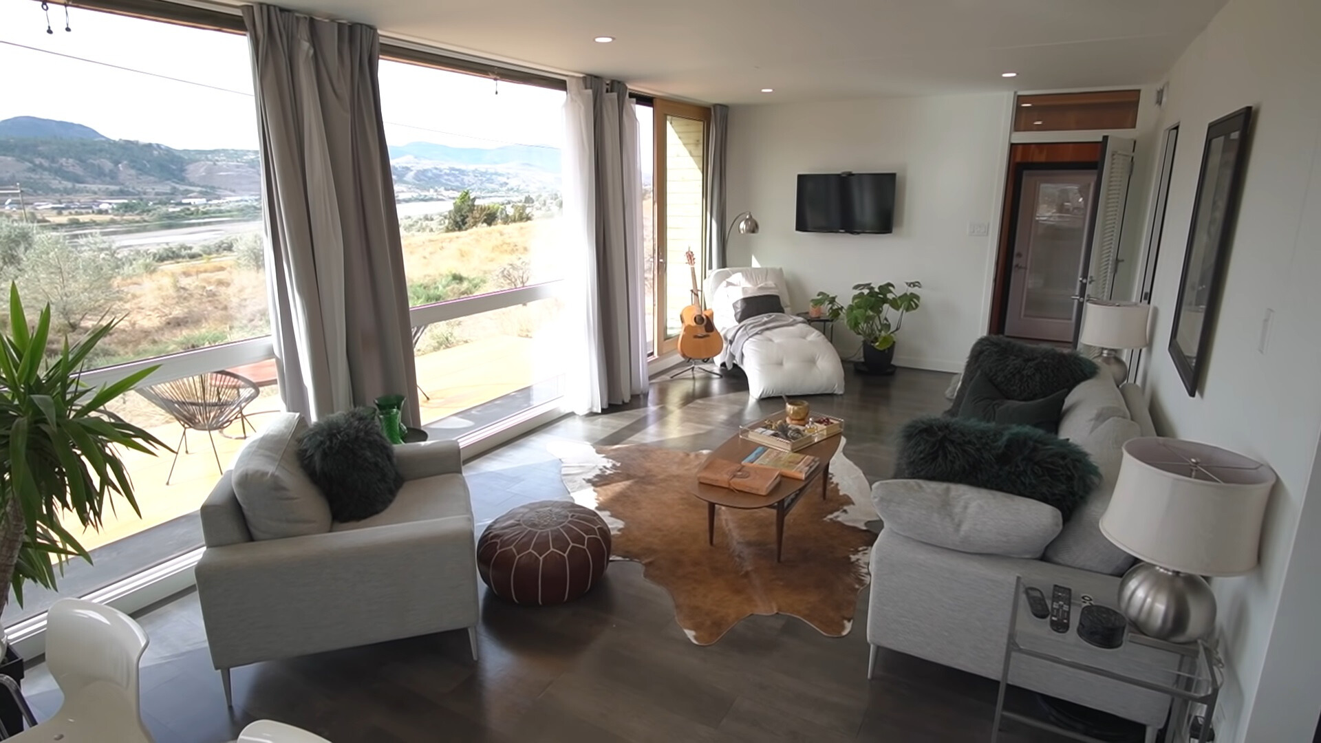 living room with floor to ceiling windows, white chair in the corner, gray couch and sofa, small brown table, cow rug