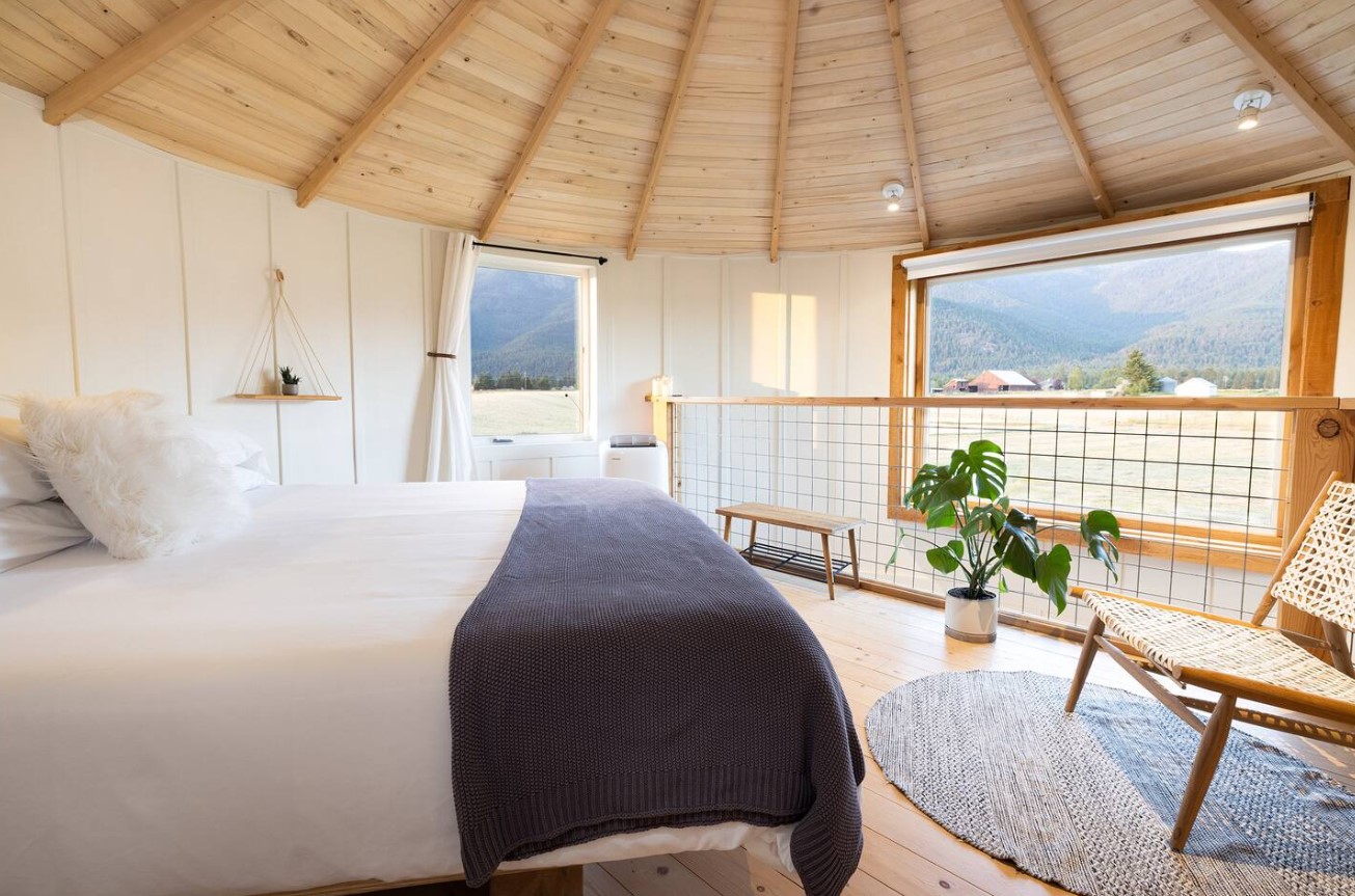 loft bedroom with a wooden ceiling, king-sized bed, chair in the corner and a small wooden bench, and a window opposite the bed