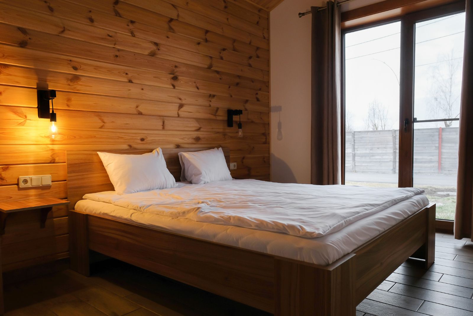 simple rustic bedroom with wood wall siding, a classic bed and white bedding, doors on the right leading out on the balcony