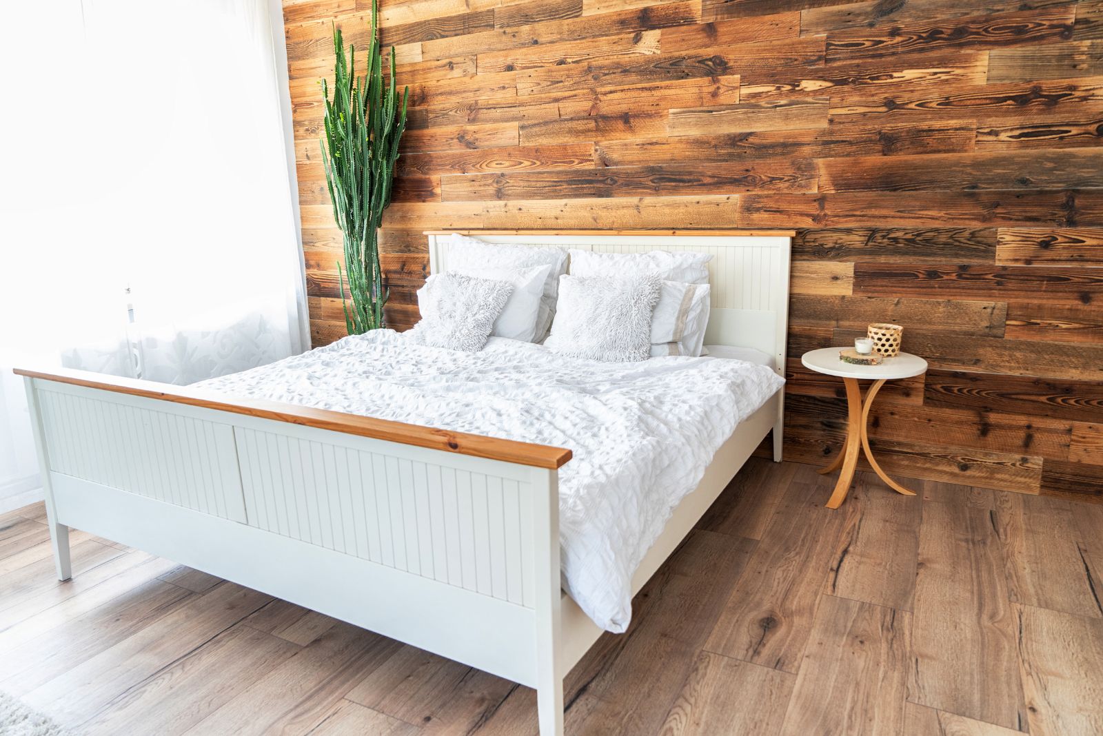 rustic bedroom with an interesting wooden accent wall behind the bed, white bed with white bedding, small side table, a plant on the left and airy curtains