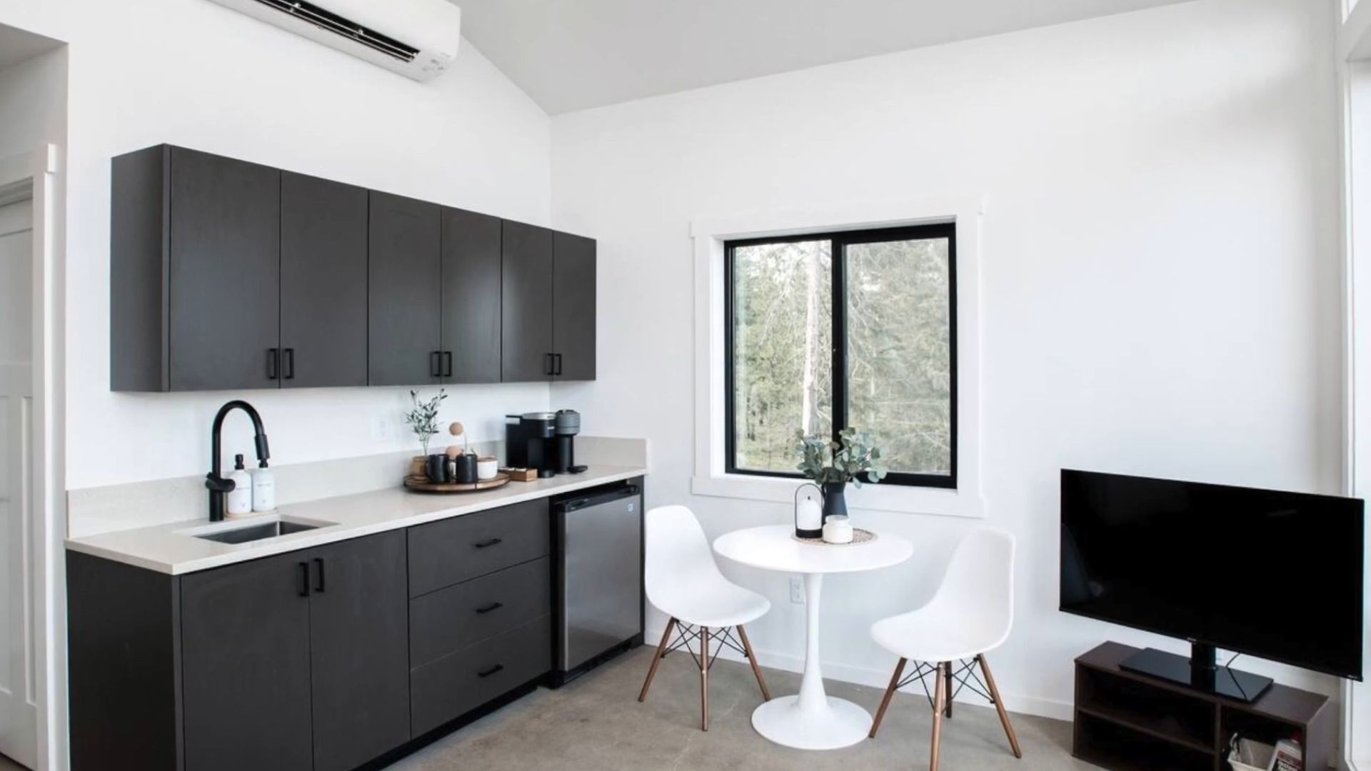 kitchen with blackish-grey cabinets, a small white dining table with two chairs next to the window, a TV in the right corner