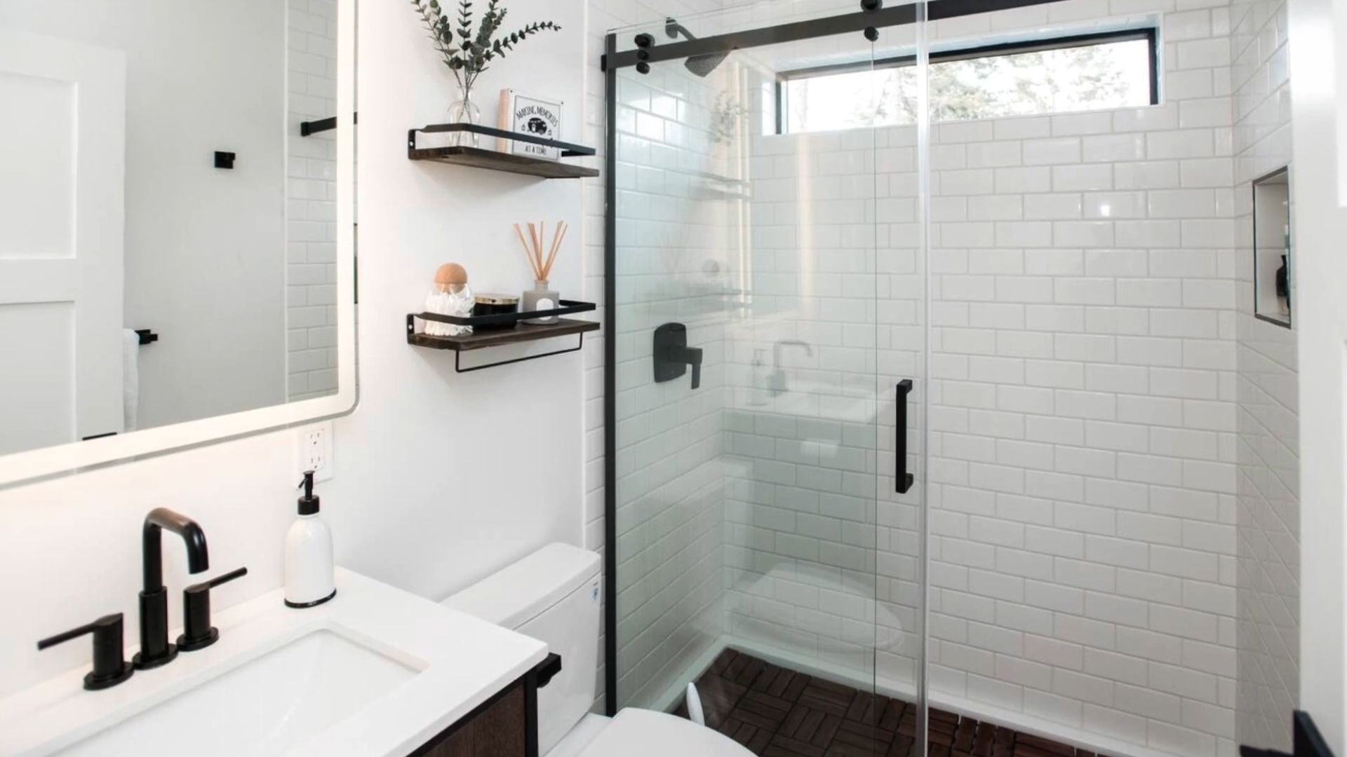 simple white bathroom with a walk-in shower, floating shelves next to it, a black and white sink with a black faucet, a bi mirror, and lots of details in black