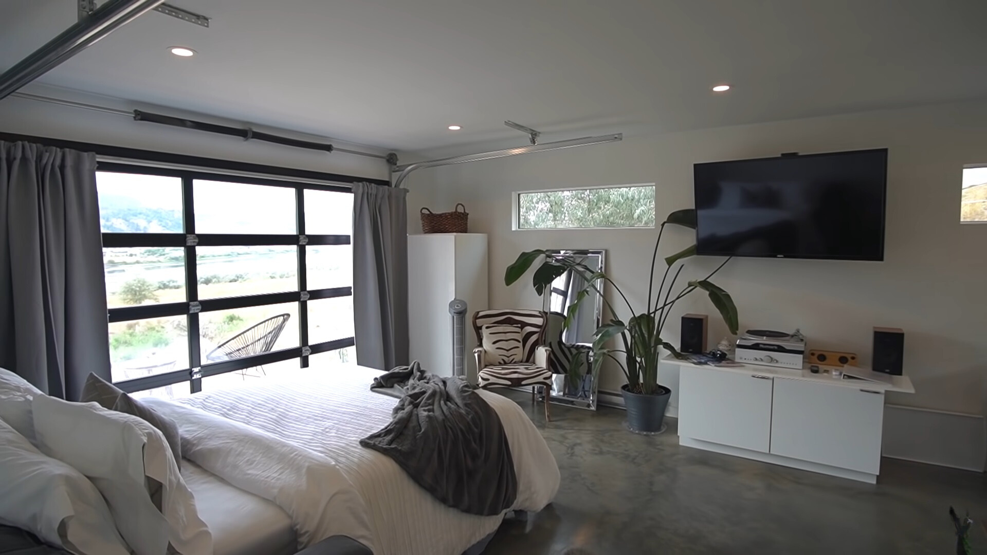 studio with a bed, doors which are also floor to ceiling windows, flat screen tv, white cabinet with a record player on top, a mirror, big plant next to the mirror and a zebra chair