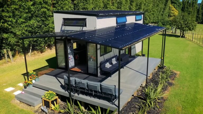 exterior of the tiny house on wheels, with a covered deck and a seating area, surrounded by forest and greenery