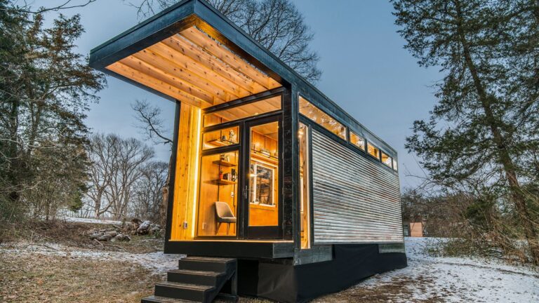 exterior of the tiny house Cornelia with wood siding, black steel frame, small stairs, lights on at night