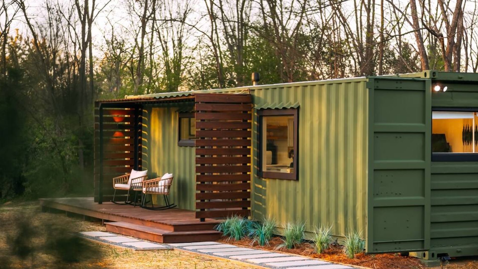 exterior of the forest green container with black frame windows and a covered porch, woods around it