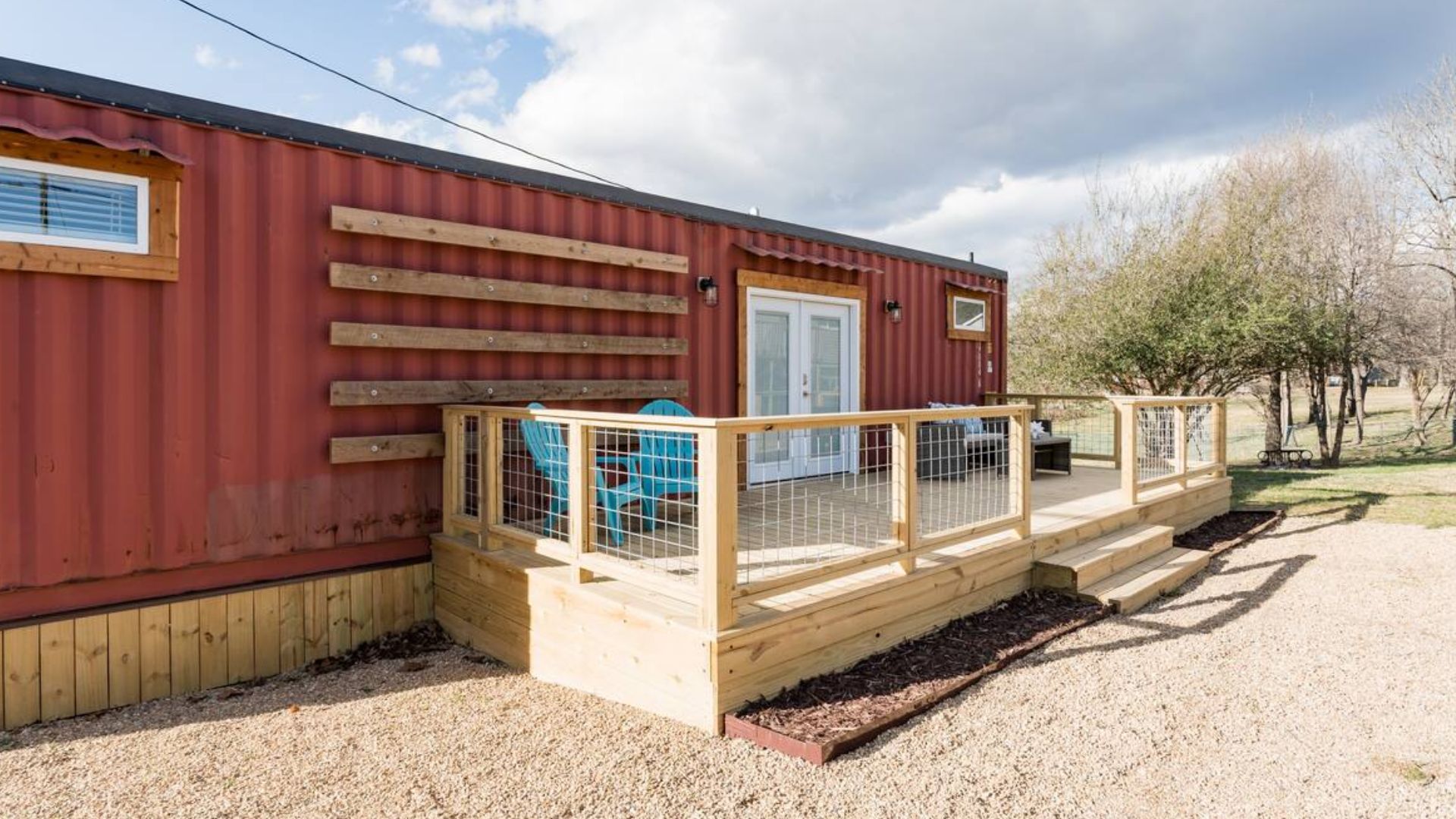 exterior of the container house with red corrugated siding, a spacious deck with a wire fence, two seating aareas, some wooden shelves on the left side of the container near the entrance