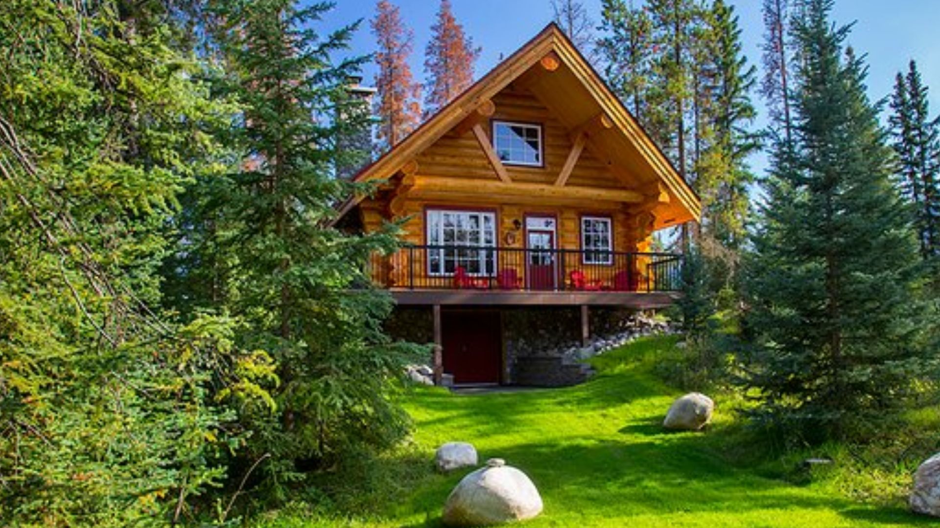 exterior of the small rustic cabin in Alberta with red frames around fenestration, a-frame red roof with a stone chimney on the right, wrap-around porch, surrounded by pine trees