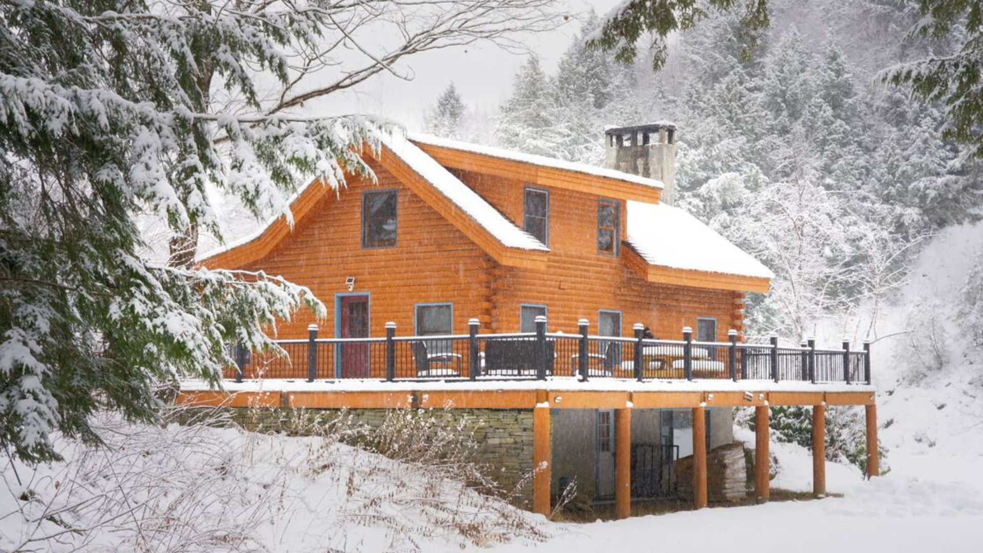 front side of the cozy log cabin in a lush forest, with a wooden porch, stone chimney, and a spacious deck, in winter