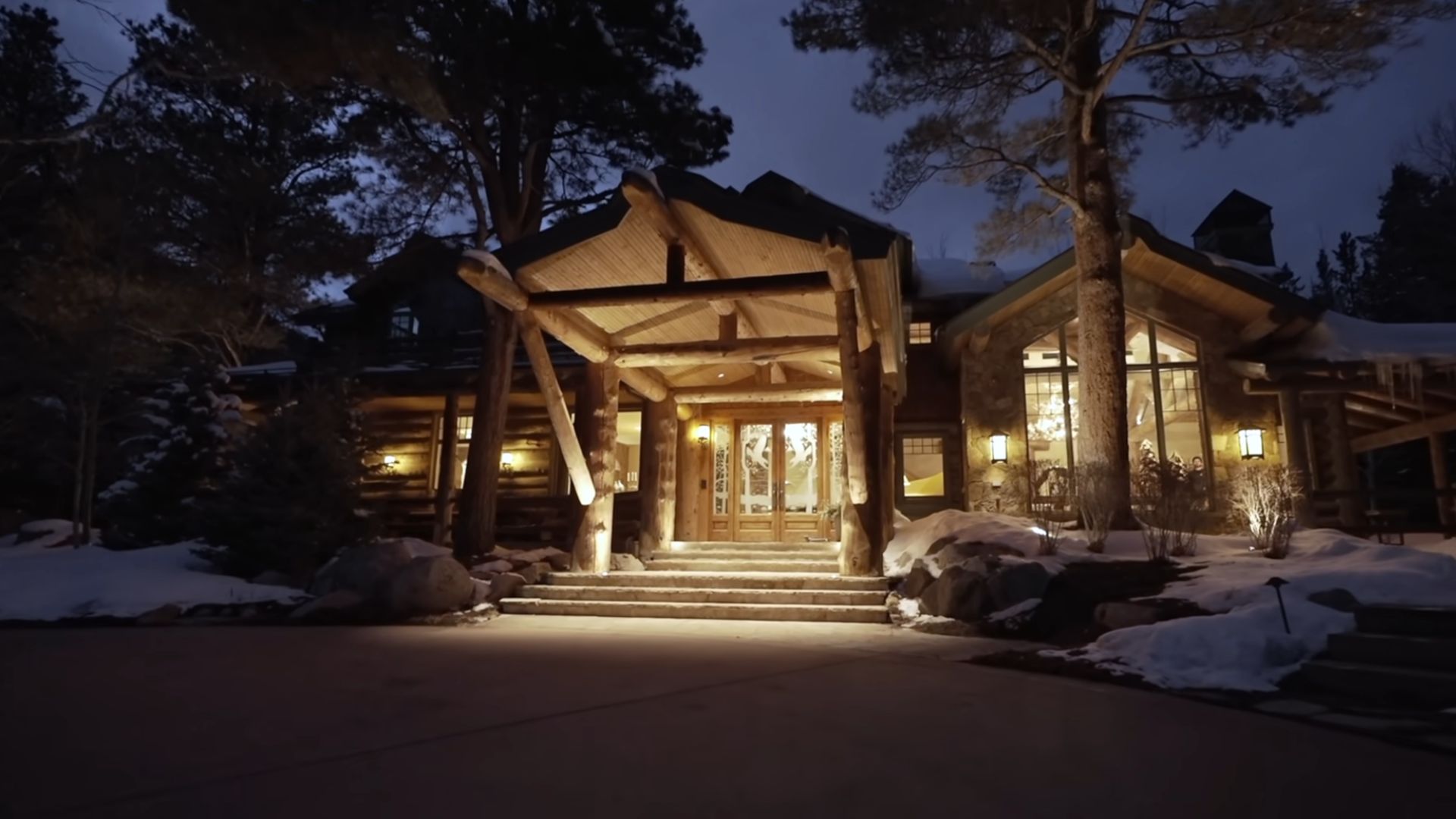 A large, rustic-style mountain cabin with a stone and wood exterior, surrounded by snow-covered trees and landscape, a steeply pitched roof, large windows, and a welcoming entrance with a wooden pergola at night