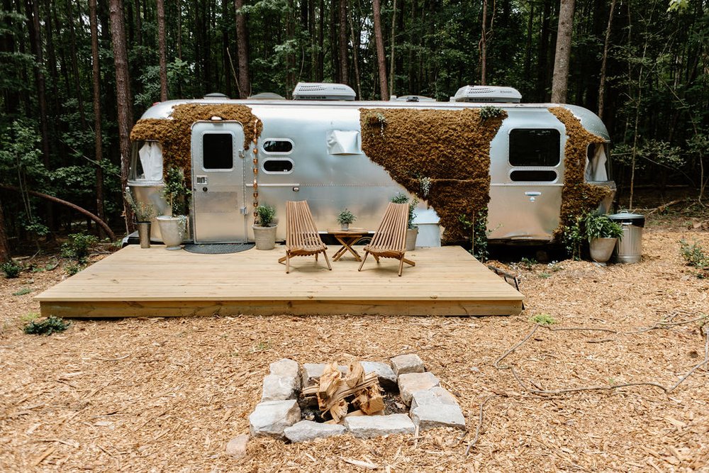 exterior of an airstream in the woods with a little terrace in front with brown chairs and a firepit