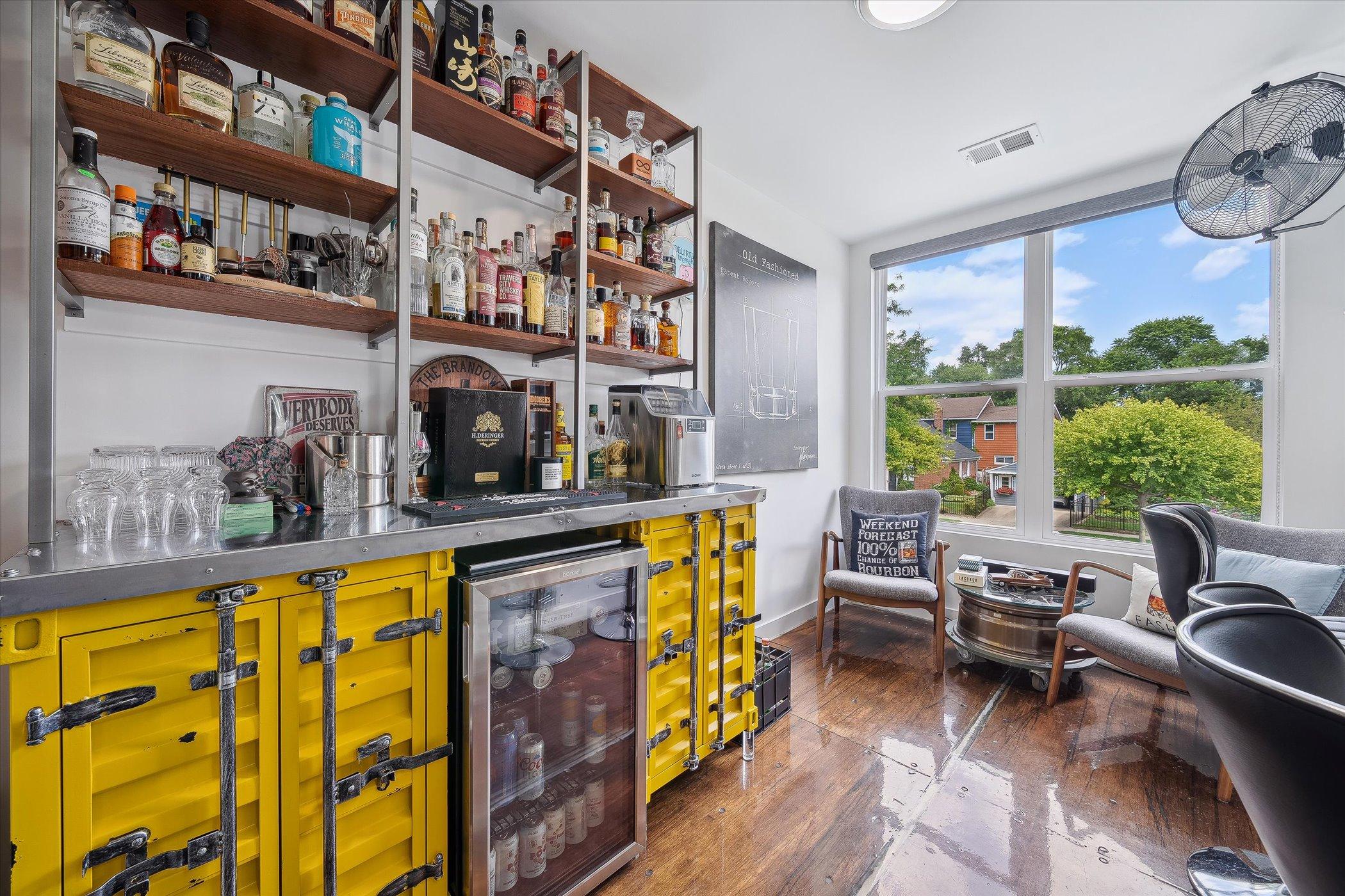 bar room with a lot of drinks on the shelves and in the mini fridge, two chairs near the window and three black bar stools