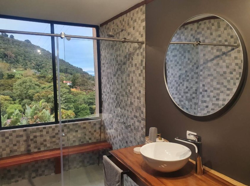 bathroom with a large window, spacious glass shower, round mirror, white sink and a brown cabinet