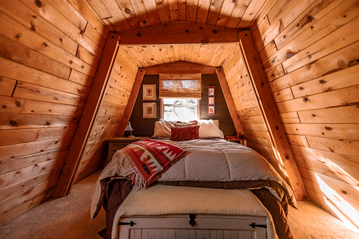 bedroom with brown, wooden walls, one bed, window above the bed, bedside tables with lamp on top, photos on the wall