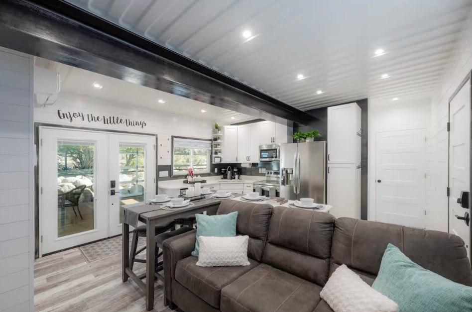 living room with white walls, brown couch with light blue and white pillows, glass doors, kitchen in the background