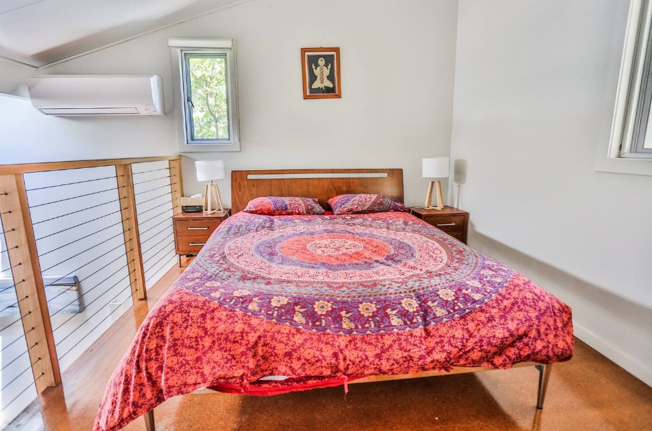 bedroom with a textured red and purple bed sheet, brown bedside tables on each side of the bed with lamps on them, window and white walls