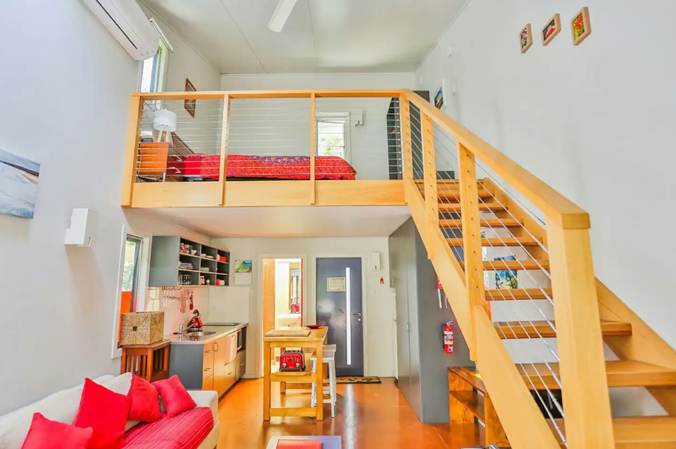 interior of a container with a living room with red pillows, kitchen with a brown island in the background and brown stairs on the right that lead up to the bedroom on the second floor