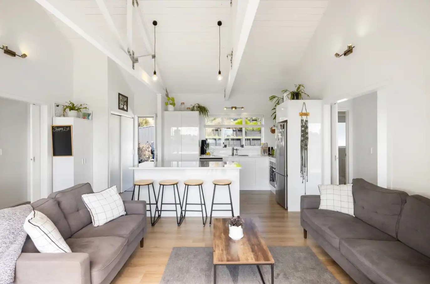 living room with gray couches, wooden table in the middle, kitchen with white cabinets and bar stools in the background