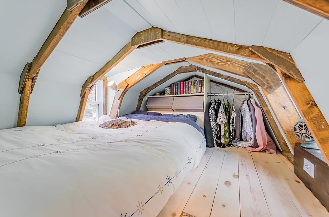 loft bedroom with white walls, comfy bed, clothes hung on one wall and a window on the side