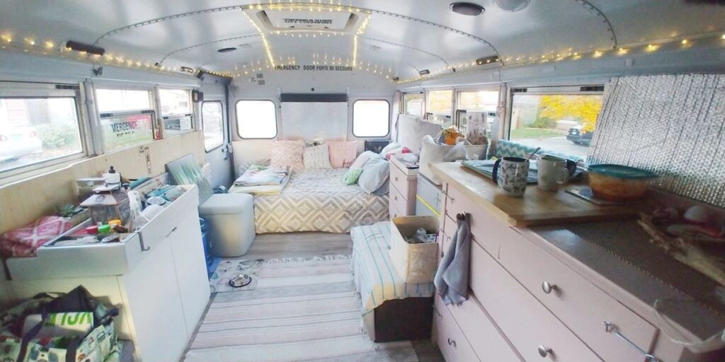 interior of a converted school bus with a comfy bed and white and light pink cabinets