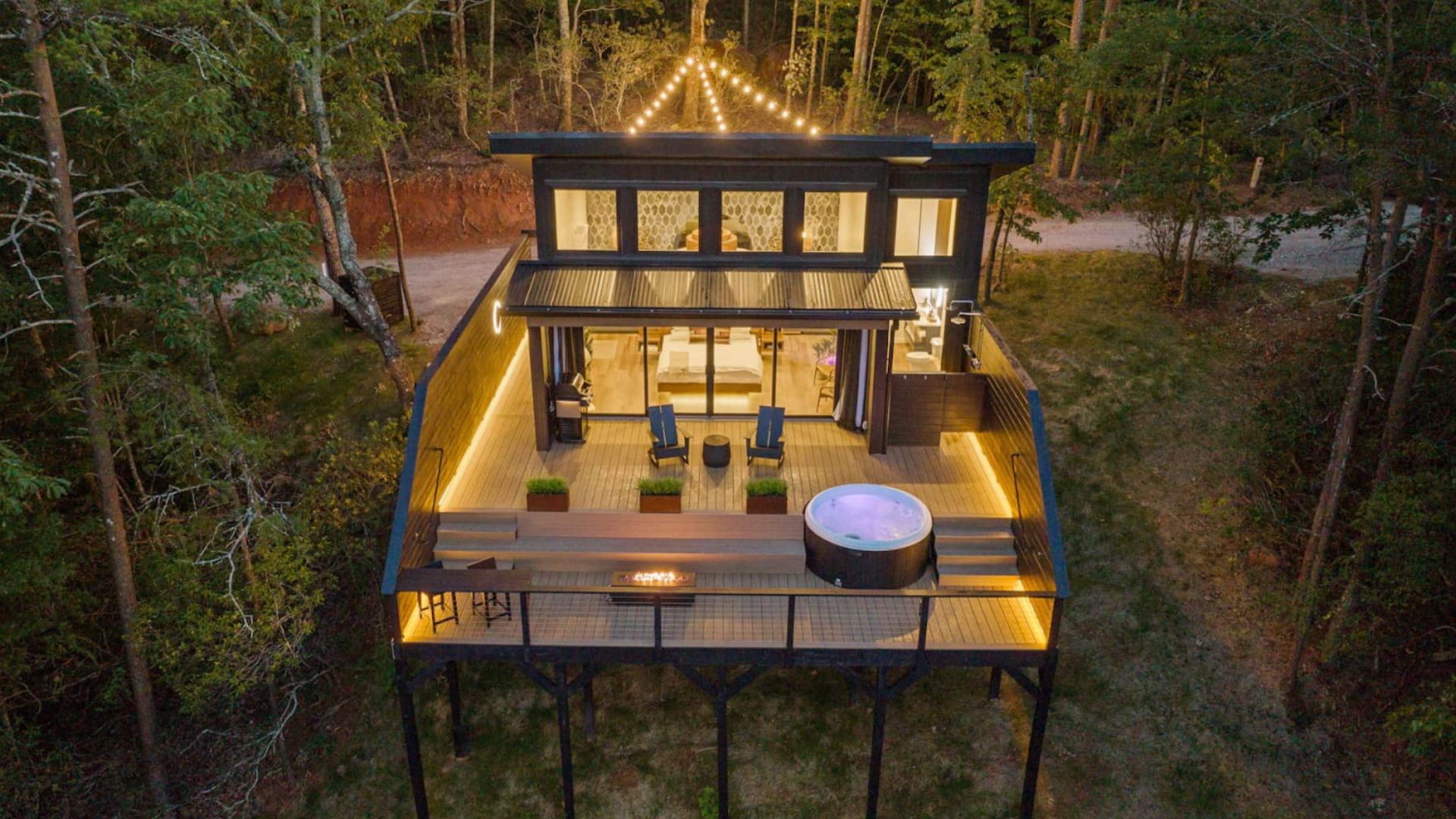 exterior of the tiny house looking like a cool cabin in the woods, huge deck, hot tub on it, chairs, lights on in the house