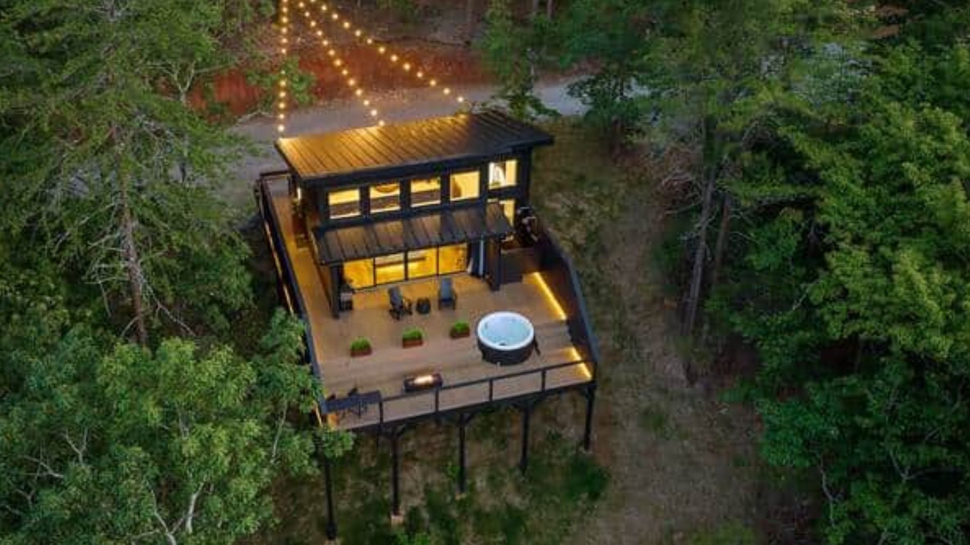 exterior of the tiny house looking like a cool cabin in the woods, huge deck, hot tub on it, chairs, lights on in the house