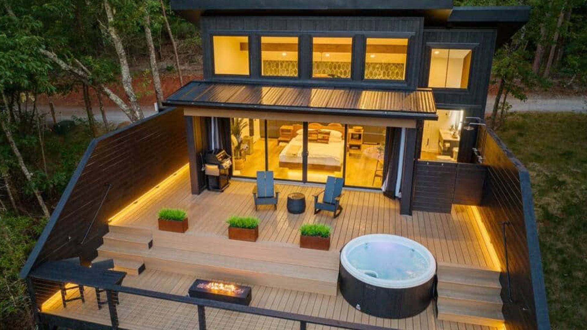 exterior of the tiny house looking like a cool cabin in the woods, huge deck, hot tub on it, chairs, lights on in the house