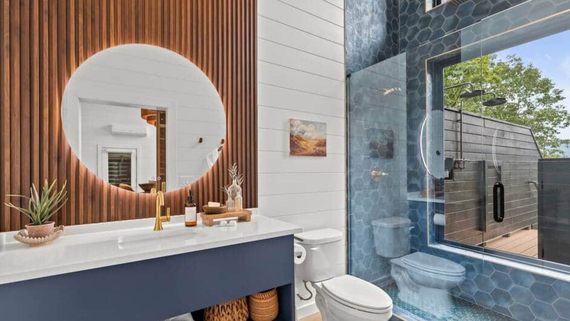 luxurious bathroom with a pretty blue walk-in shower, blue hexagonal tiles, blue floatingvanity, a round mirror, wood details behind the mirror, and golden accents