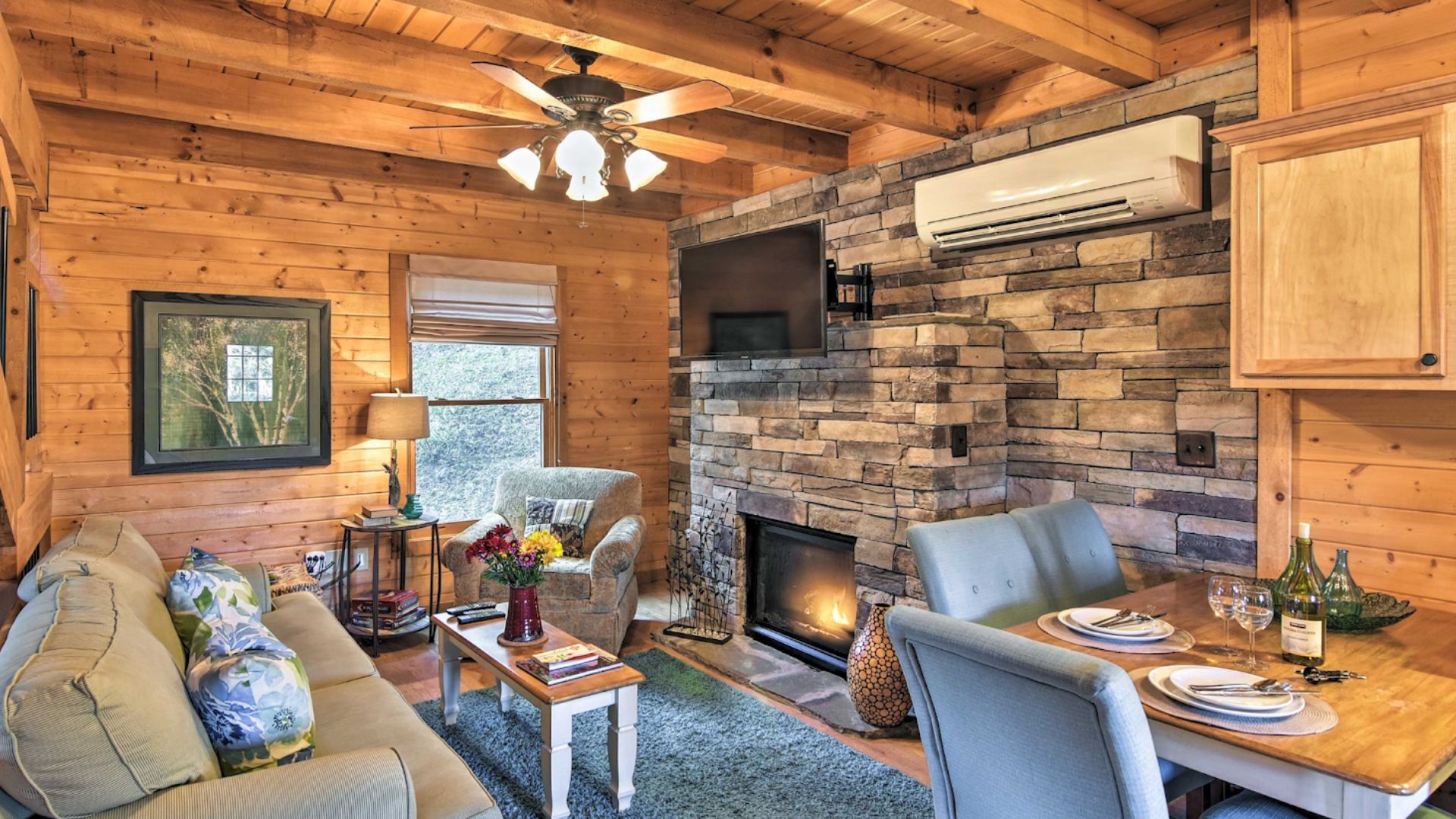 living room with grey sofa, an accent wall in grey stone with a TV on it and a modern fireplace unde the TV, dining room chairs on the right and a kitchen cabinet