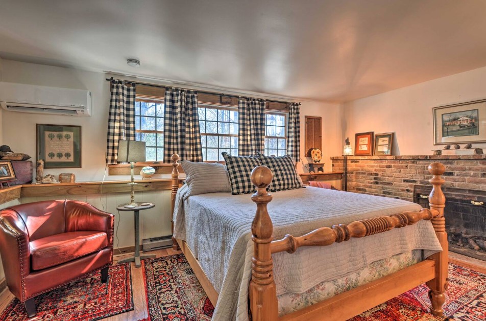 bedroom with a big bed, checkered curtains over the windows, dark red comfy sofa, white ac, framed photos, round bedside table