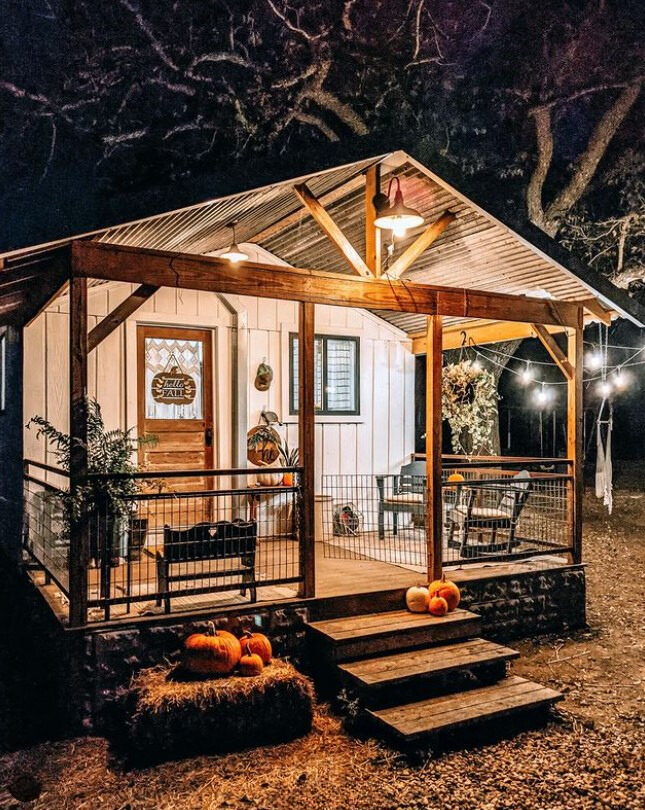 exterior of a cabin with white wall, tiny porch with two chairs and warm lightning, decorated with pumpkins and plants