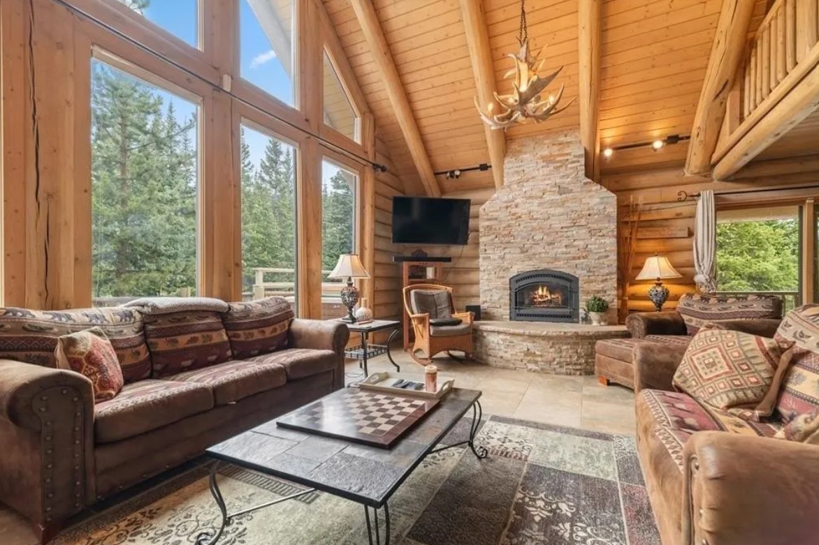 living room with floor to ceiling windows, wooden walls, beams above, brown couches, stone fireplace, flat screen tv