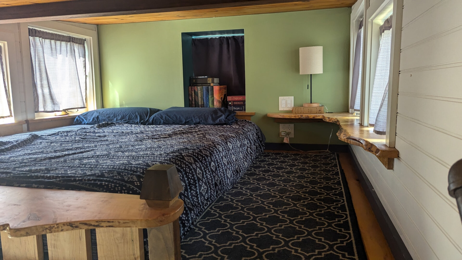 loft bedroom with a bed with dark blue bed sheets, wooden desk along the windows