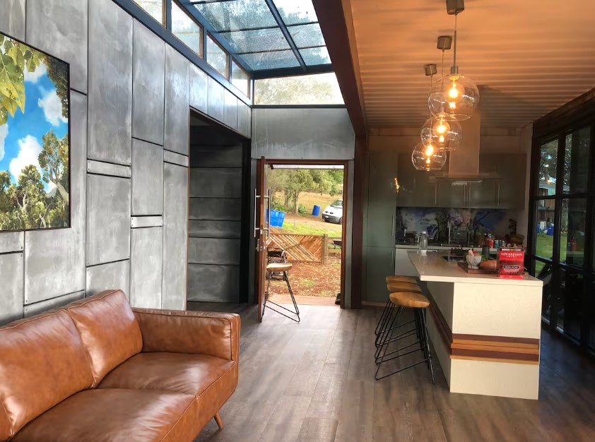 living room with a brown leather couch, kitchen in the back, bar stools under pendant lightning