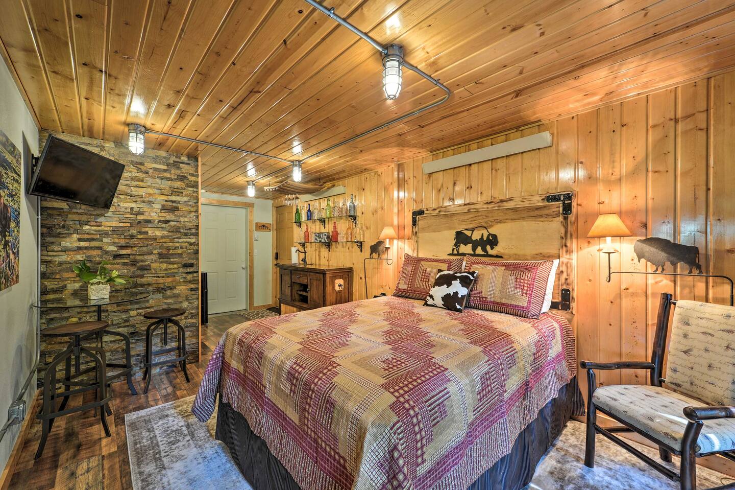 bedroom with a cozy bed, wooden walls, one stone accent wall, mini bar inside of a room with a round table in the corner, flat screen mounted tv