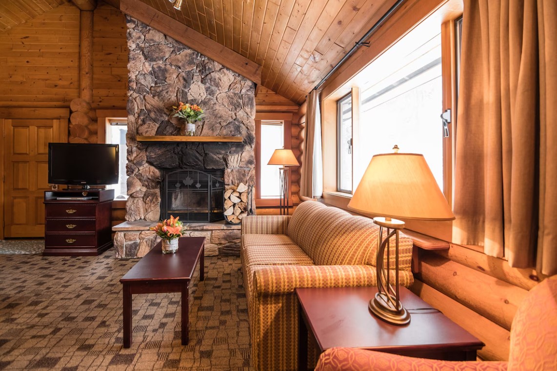 living room with a patterned couch, large window, tv, stone fireplace, brown table